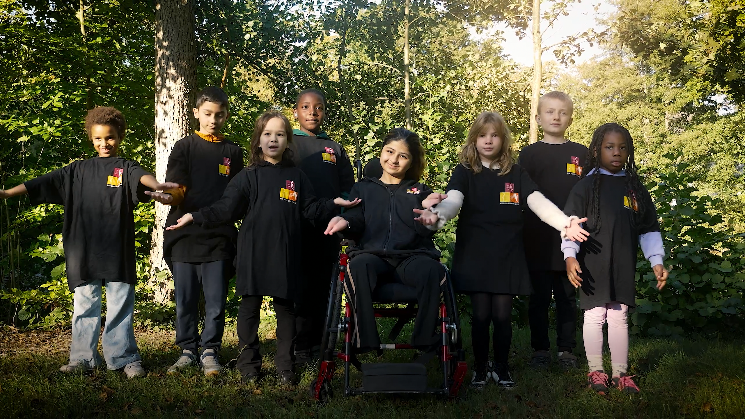 Das Bild zeigt: 8 Kinder in schwarzen T-Shirts mit Schullogo stehen im Park und singen und gebärden den Song "Hey du"