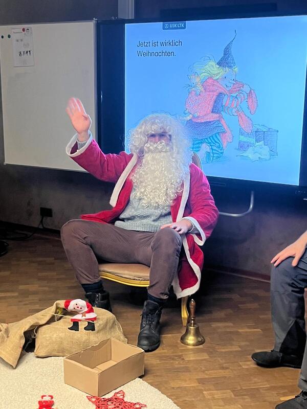 Das Bild zeit: Der Nikolaus mit einem roten Mantel und einem weißen Bart sitzt vor dem Smartboard und winkt den Kindern zu.