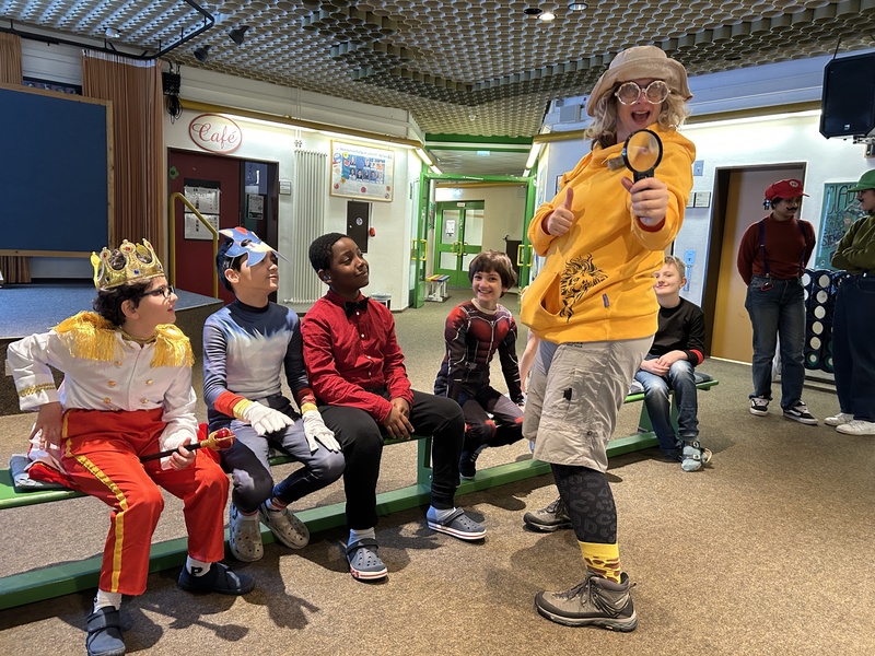 Das Bild zeigt: Eine Lehrerin, als Naturforscherin verkleidet, untersucht und erforscht mit einer großesn Lupe einige verkleidete Kinder.